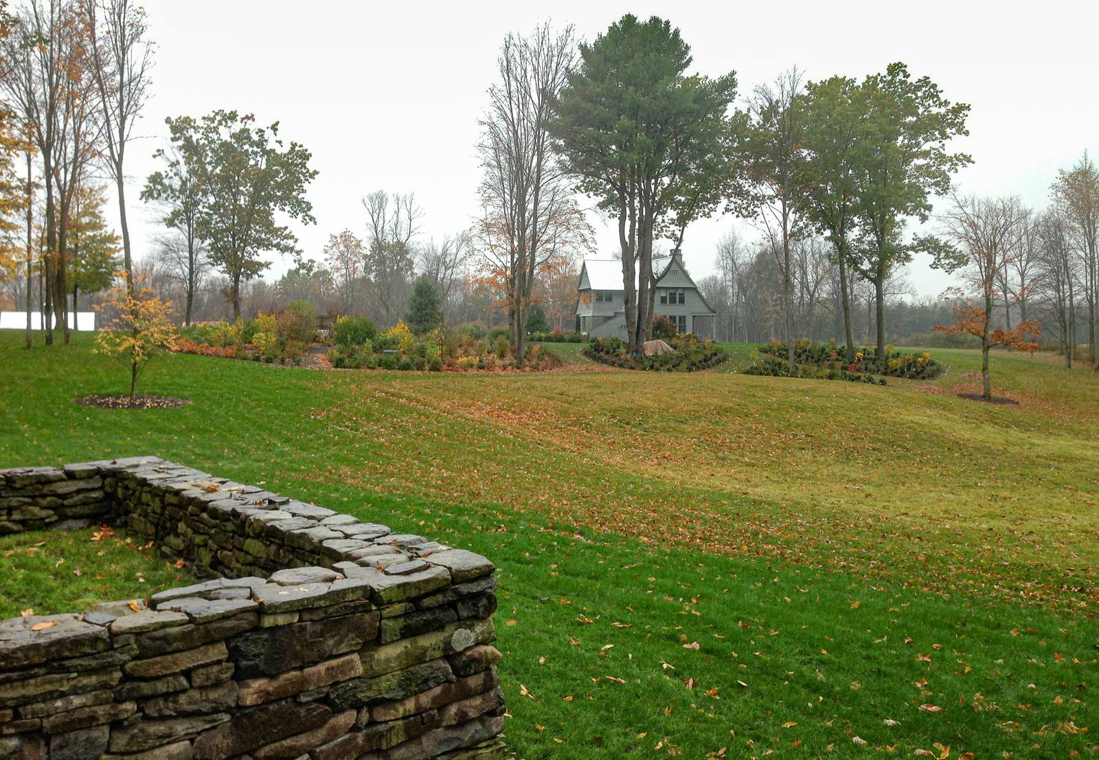 Stone wall corner above a sloping lawn, with the house and new plantings beyond
