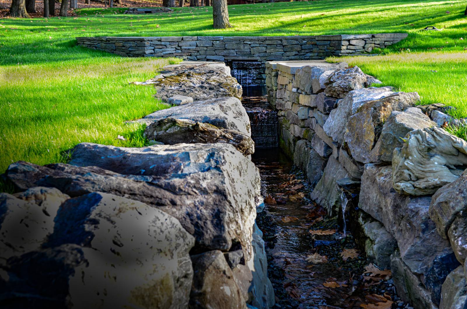 Stone-lined spring channel with a small waterfall beneath a low stone wall
