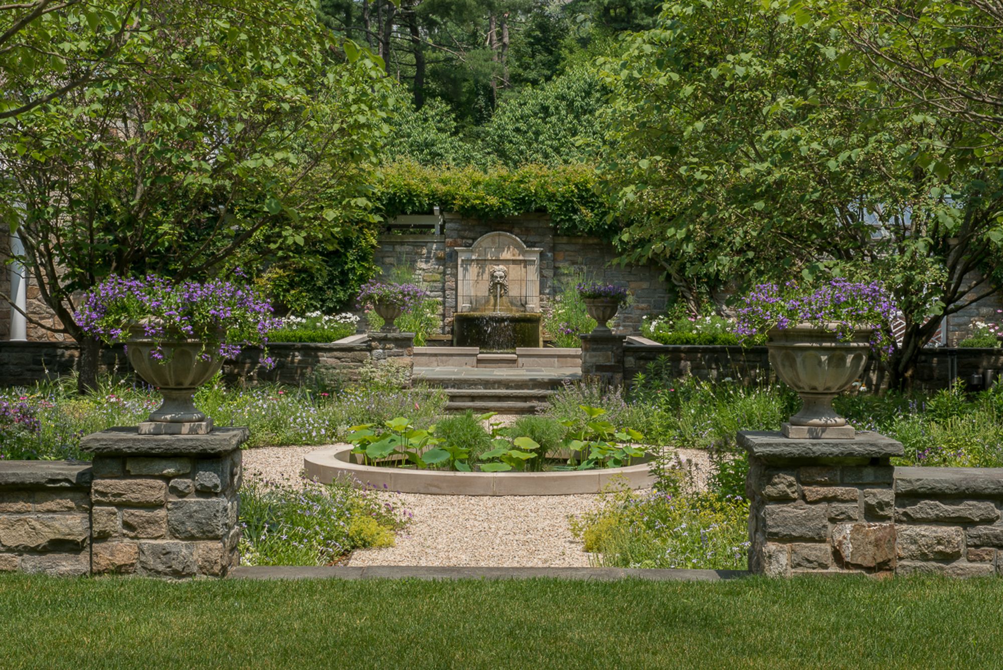 Walled garden with a lion-mask wall fountain, stone urns of purple flowers on piers and a round lotus pool in gravel