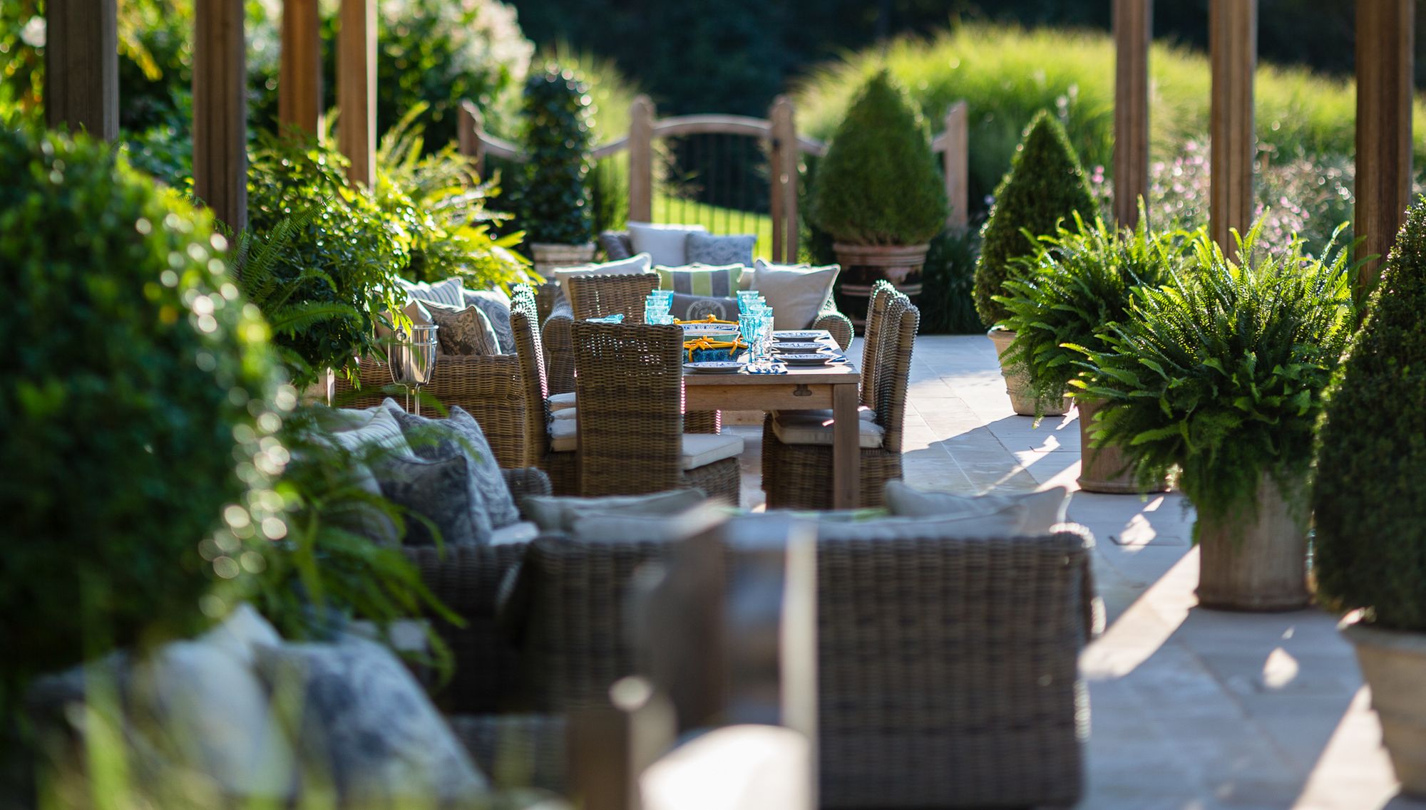 Wicker dining table set beneath a long columned pergola, lined with potted ferns and boxwood cones