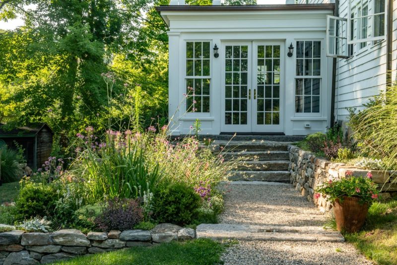 Granite steps leading to the white conservatory doors beside a dry-laid stone wall and a flowering border
