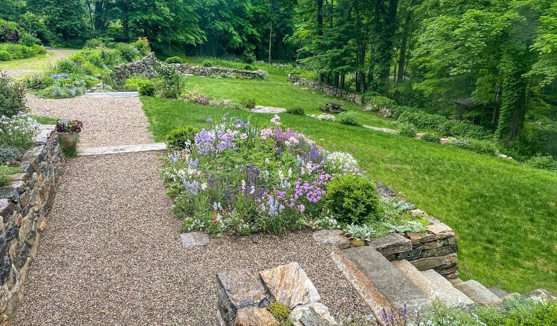 Gravel path, stone retaining wall and a perennial border stepping down to the lawn and woods
