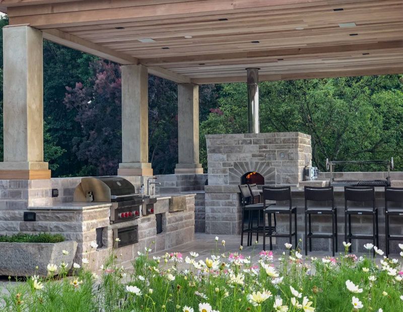 Outdoor kitchen under a cedar-ceilinged pavilion with a gas grill, limestone pizza oven and parrilla beyond