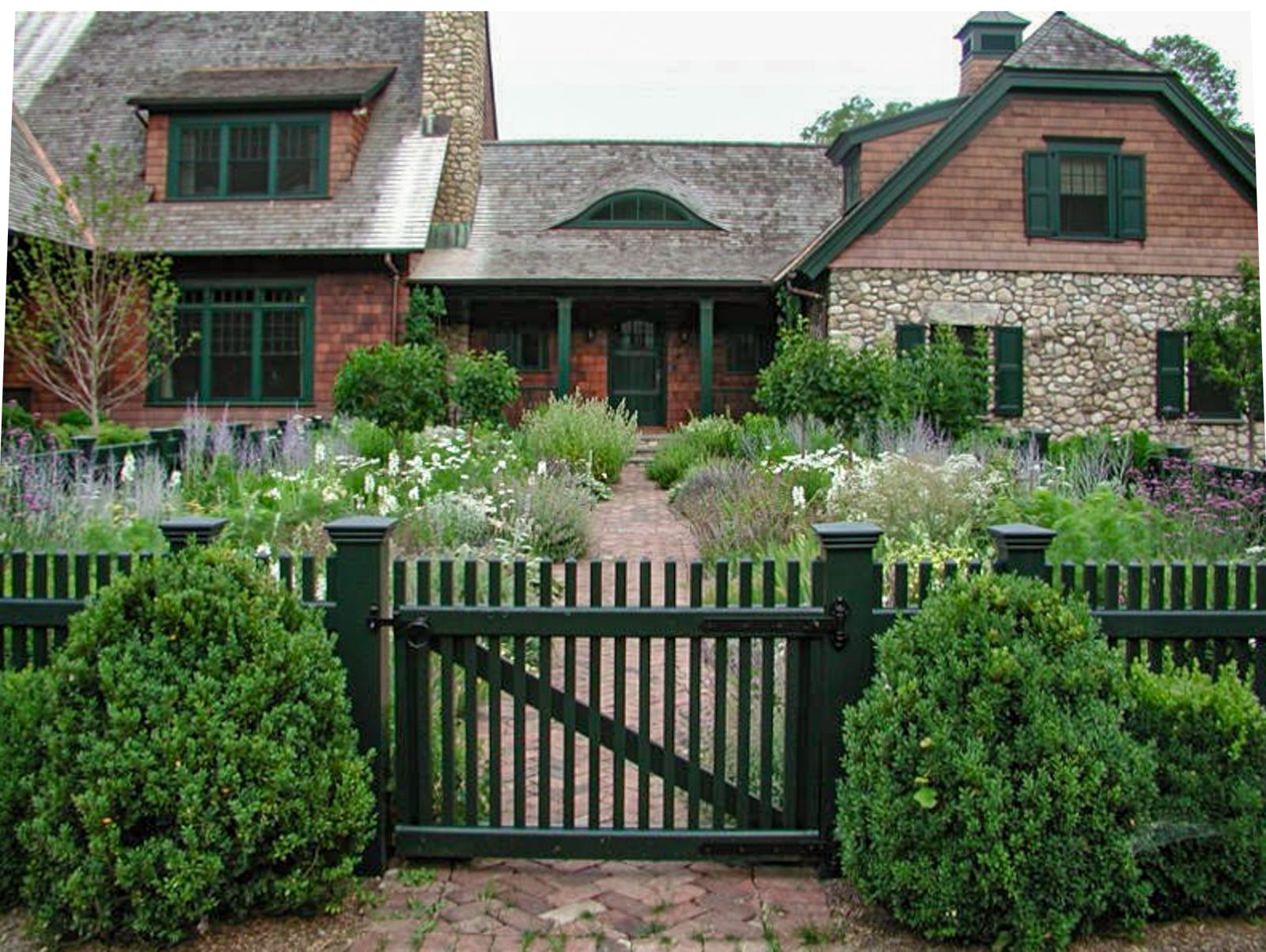 Green picket fence and gate between boxwoods opening onto a herringbone brick path through a cottage garden of lavender, catmint and white flowers before a shingle and stone house