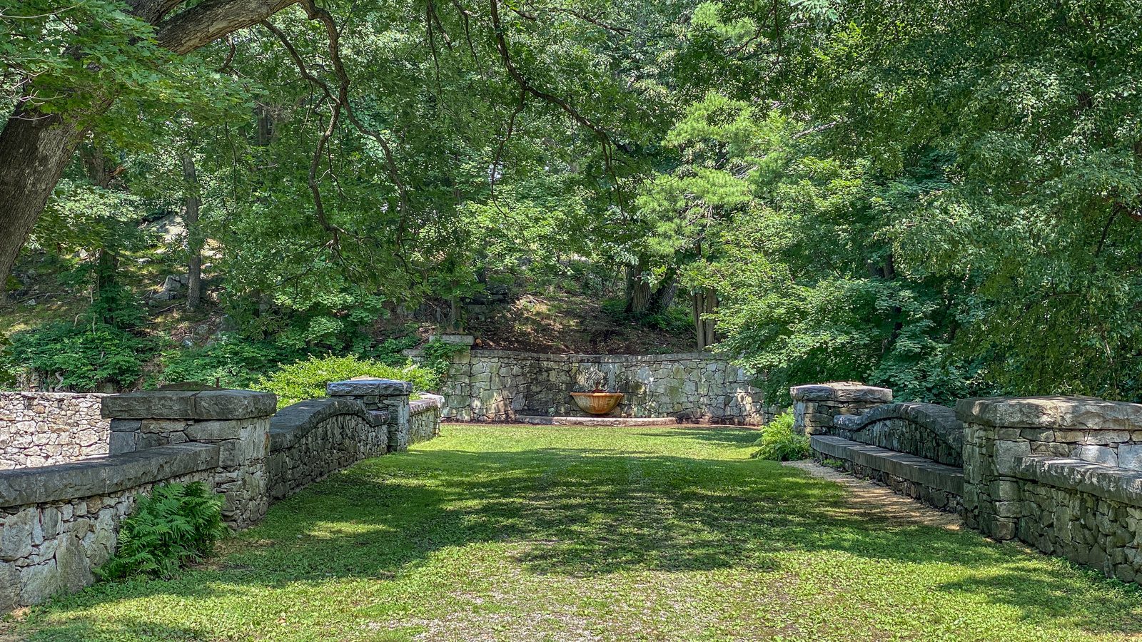 Lawn allée between stone walls and benches leading to a wall fountain
